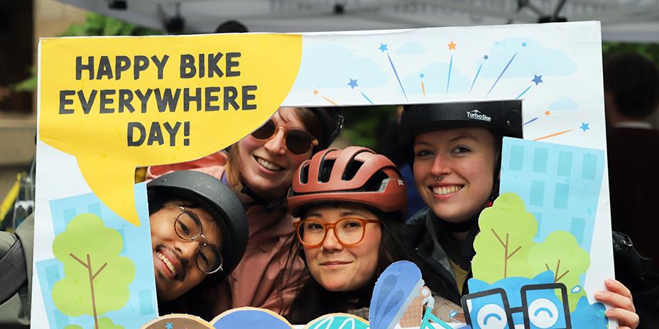 Four bicyclists pose with an illustrated frame with text, "Happy Bike Everywhere Day!"