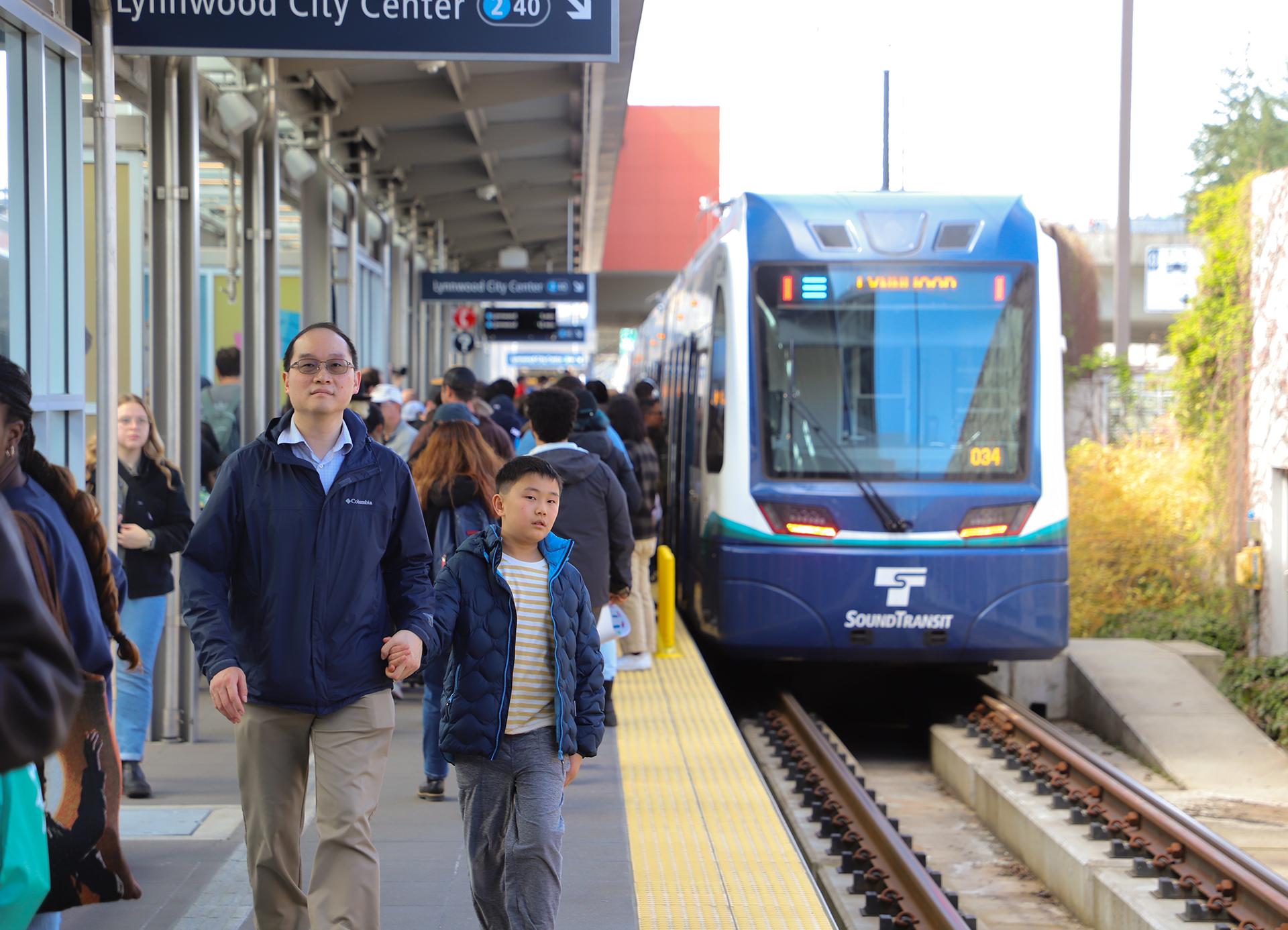 Man and boy walk along light rail station platform as riders exit a train in the background.